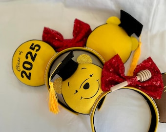 Pode incluir: Orelhas de Minnie Mouse amarelas com um chapéu de formatura do Winnie the Pooh e um laço vermelho com um charme de pote de mel. A orelha esquerda tem o texto "Class of 2025".
