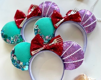 Puede incluir: Orejas de Minnie Mouse de color morado y turquesa con un lazo rojo y un dije de tenedor. Las orejas están decoradas con lentejuelas y perlas.