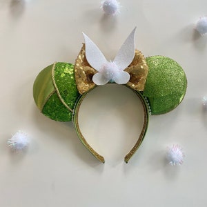 May include: Green and gold Minnie Mouse ears with a white fairy wings bow. The ears are covered in sequins and have a green satin bow.