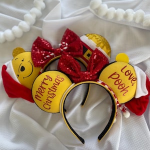 May include: Festive yellow and red Christmas headbands with Winnie the Pooh ears. One headband says "Merry Christmas" and another says "Love, Pooh". Each headband has a red sequined bow and a Santa hat.