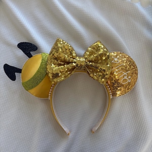 May include: A yellow headband with bee-themed ears and a sequined bow. The ears are yellow with gold sequins and the text "XIX". The bow is gold sequined. The headband has a white interior and a yellow exterior.