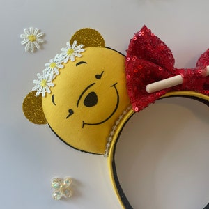 May include: Yellow Winnie the Pooh character headband with a red sequined bow and white daisy embellishments. The headband is black with a white pearl trim.