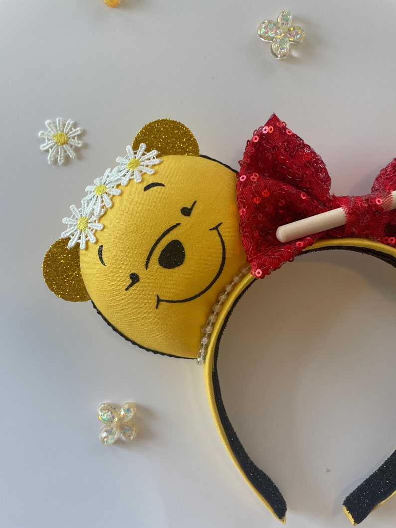 May include: Yellow Winnie the Pooh character headband with a red sequined bow and white daisy embellishments. The headband is black with a white pearl trim.