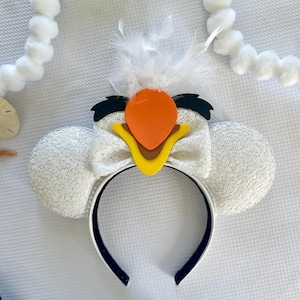 May include: White sequined mouse ear headband with a bow, featuring a cartoon bird design. The bird has an orange beak, yellow accents, and black details. White feathers are attached to the top of the headband.