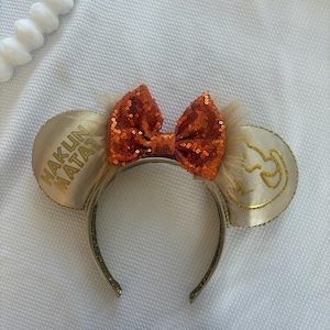 May include: A gold headband with two round ears and an orange sequined bow. The left ear features the words "Hakuna Matata" in gold, and the right ear has a gold lion design.