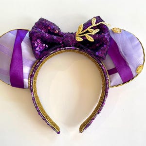 Puede incluir: Orejas de Minnie Mouse moradas y doradas con un lazo de lentejuelas y un dise&ntilde;o de hoja de oro. Las orejas est&aacute;n hechas de una tela suave y de felpa y tienen una cinta morada.