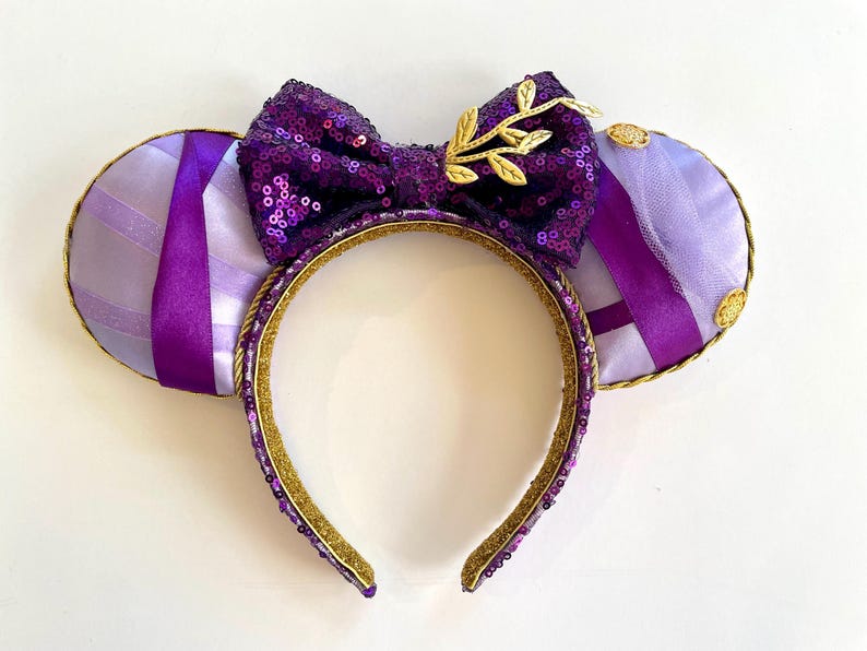 Puede incluir: Orejas de Minnie Mouse moradas y doradas con un lazo de lentejuelas y un dise&ntilde;o de hoja de oro. Las orejas est&aacute;n hechas de una tela suave y de felpa y tienen una cinta morada.
