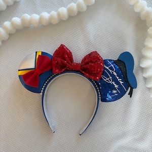 May include: Blue and white headband with sequined ears and a red sequined bow. One ear features a yellow and red design, the other has the text "Donald Duck #1" in white script. The headband is trimmed with white beads.