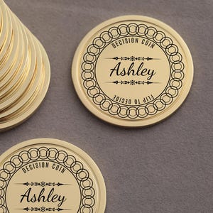 May include: A stack of gold-colored decision coins with the text "Decision Coin" and "Ashley" engraved on the front. The coins are arranged on a gray surface.