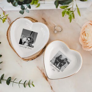 May include: Two heart-shaped trinket dishes with gold rims. One dish rests on a wooden base. Each dish features a black and white photo and text. One says "Soul Sister Forever", the other says "Claire Charlotte 2025". A silver ring is also present.
