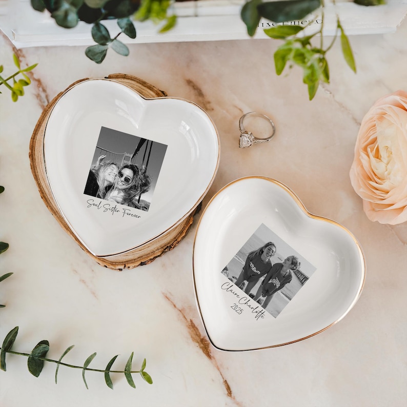 May include: Two heart-shaped trinket dishes with gold rims. One dish rests on a wooden base. Each dish features a black and white photo and text. One says "Soul Sister Forever", the other says "Claire Charlotte 2025". A silver ring is also present.