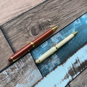 May include: Two pens with gold accents on a woodgrain background. One pen is brown wood and the other is light wood.