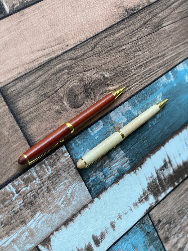 May include: Two pens with gold accents on a woodgrain background. One pen is brown wood and the other is light wood.
