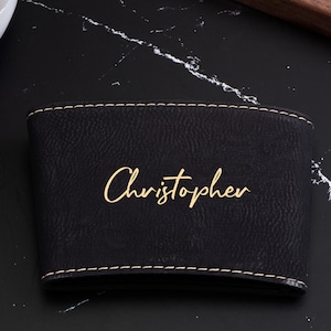 Puede incluir: Funda para taza de caf&eacute; de cuero negro con un borde cosido dorado y el nombre "Christopher" escrito en letras doradas.