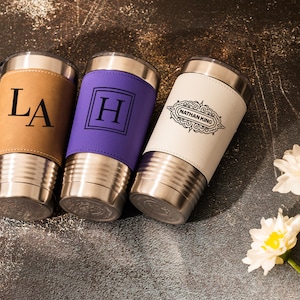 May include: Three stainless steel tumblers with personalized leather wraps. The tumblers are brown, purple, and white. The brown tumbler has the initials "LA" engraved on it. The purple tumbler has the letter "H" engraved on it. The white tumbler has the text "Nathan King" engraved on it.