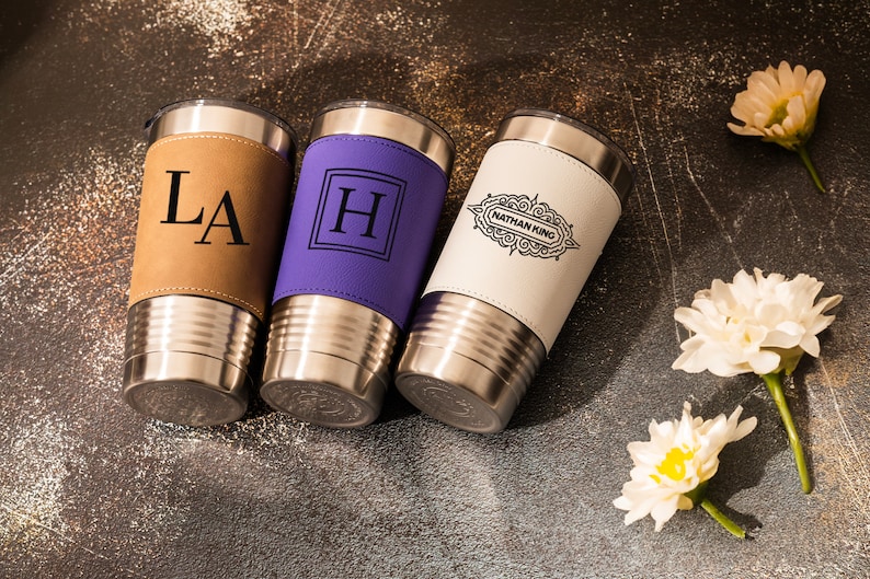 May include: Three stainless steel tumblers with personalized leather wraps. The tumblers are brown, purple, and white. The brown tumbler has the initials "LA" engraved on it. The purple tumbler has the letter "H" engraved on it. The white tumbler has the text "Nathan King" engraved on it.