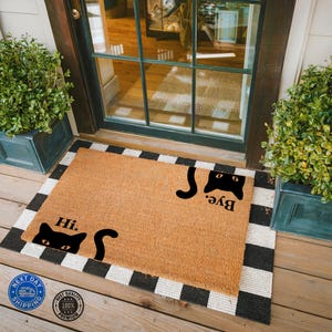 May include: A brown coir doormat with black and white checkered border. The doormat has two black cat silhouettes with the words "Hi." and "Bye." printed on it.
