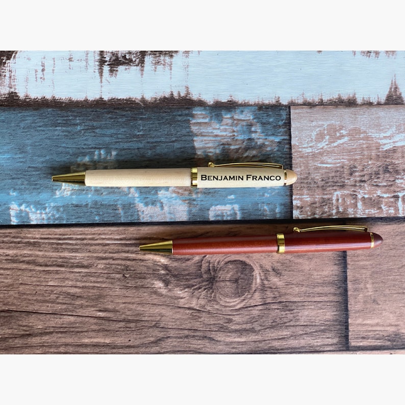 May include: Two wooden pens with gold accents. The top pen is light wood with the name "Benjamin Franco" engraved on it. The bottom pen is dark wood.