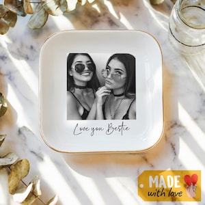 May include: A white square ceramic trinket dish with a gold rim. The dish features a black and white photo of two women wearing sunglasses, with the text "Love you Bestie".