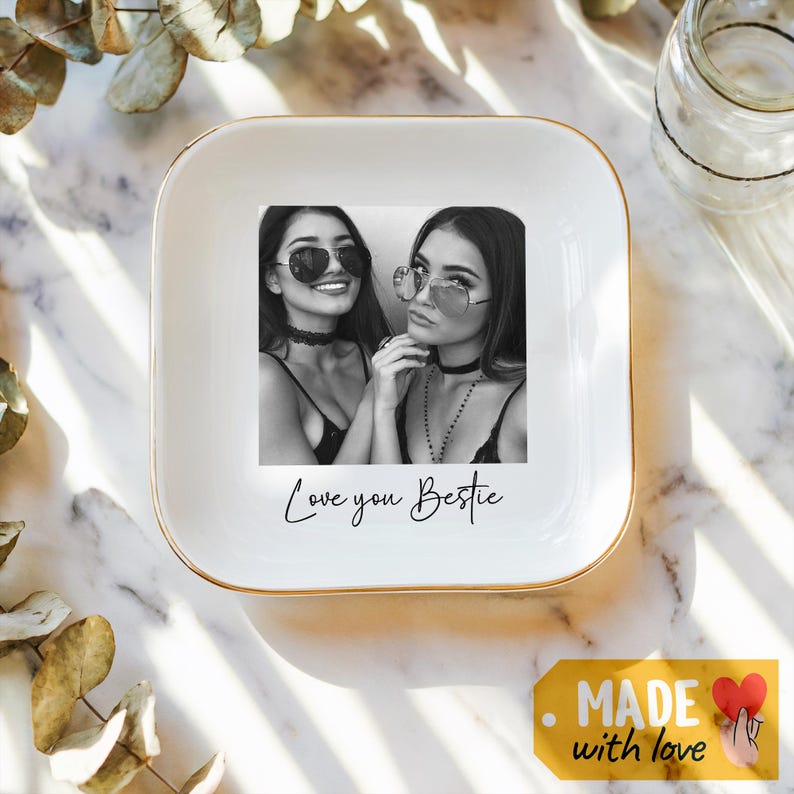 May include: A white square ceramic trinket dish with a gold rim. The dish features a black and white photo of two women wearing sunglasses, with the text "Love you Bestie".