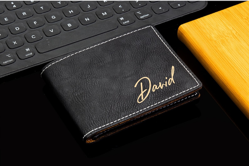 Puede incluir: Una cartera de cuero negro con costuras blancas y una inscripci&oacute;n dorada que dice "David".