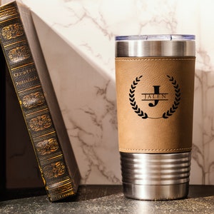 May include: A stainless steel travel mug with a brown leatherette wrap. The mug has a black engraved design with the letter "J" and the name "Jalen" in a wreath.