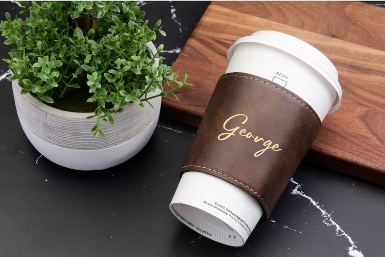 Puede incluir: Una funda de taza de caf&eacute; de cuero marr&oacute;n con el nombre "George" grabado en letras doradas. La funda est&aacute; en una taza de caf&eacute; de papel blanca.