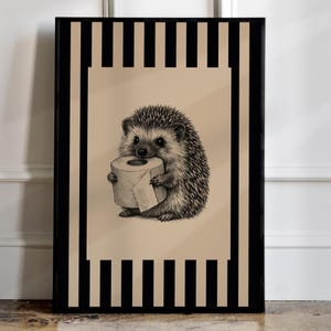 May include: A framed wall art print featuring a detailed black and white illustration of a hedgehog holding a roll of toilet paper. The central image is set against a tan background, framed by a border of vertical black and tan stripes.
