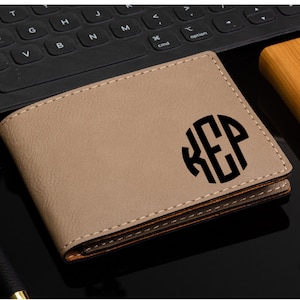 Puede incluir: Una cartera de cuero marr&oacute;n con un borde cosido marr&oacute;n. La cartera tiene un monograma circular negro con las letras "K.E.P."