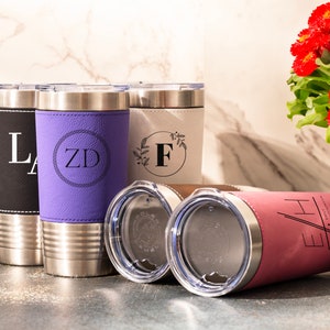 May include: Four stainless steel tumblers with different colored leather wraps and personalized monograms. The tumblers are on a light gray countertop. The tumblers are black, purple, white, and pink.