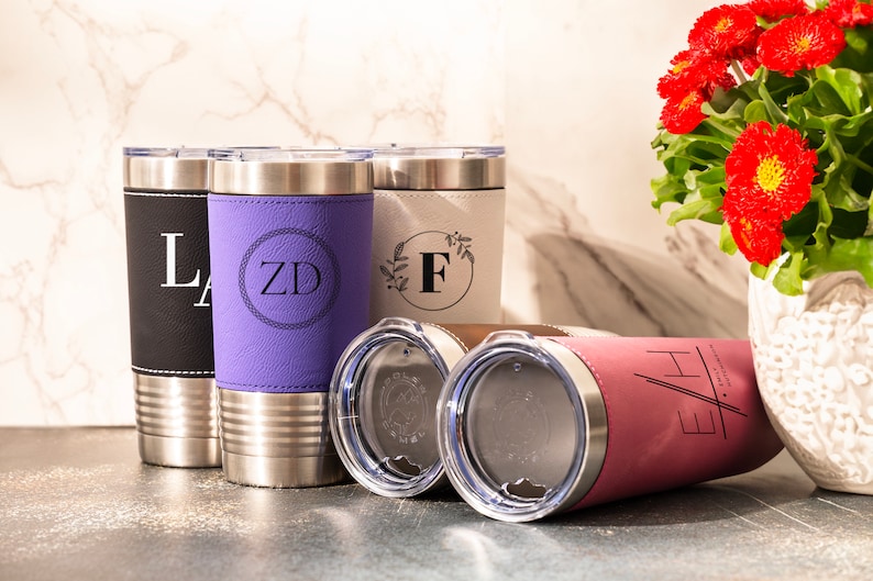 May include: Four stainless steel tumblers with different colored leather wraps and personalized monograms. The tumblers are on a light gray countertop. The tumblers are black, purple, white, and pink.