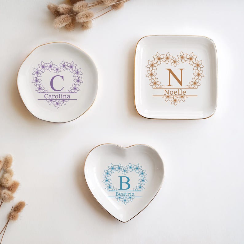 Op de afbeelding: Drie witte keramische sieradenbakjes met gouden randen. E&eacute;n rond, &eacute;&eacute;n vierkant en &eacute;&eacute;n hartvormig. Elk bakje heeft een bloemenkransontwerp met een letter en naam: "C Carolina", "N Noelle" en "B Beatriz".