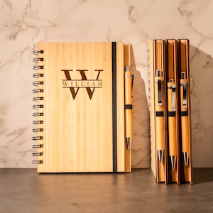 May include: A wooden journal with a spiral binding and a personalized engraved initial "W" with the name "William" below. The journal is next to three wooden pens with silver accents.