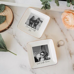 May include: Two square, white ceramic trinket dishes with gold trim. Each dish features a black and white photograph. One says "Claire Charlotte 2025" and the other says "Soul Sister Forever". A ring with a small stone sits nearby.