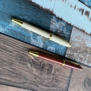 May include: Two wooden pens with gold accents. The pen on the left is light wood and the pen on the right is dark wood.