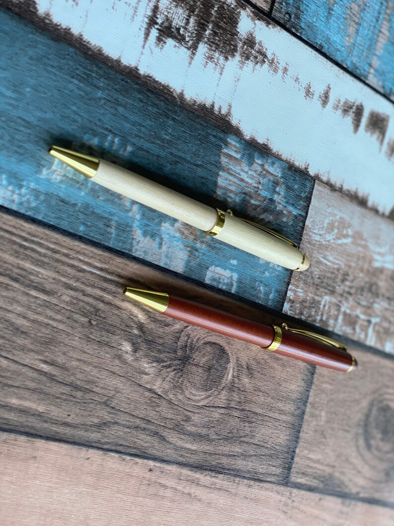 May include: Two wooden pens with gold accents. The pen on the left is light wood and the pen on the right is dark wood.