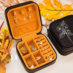 Puede incluir: Caja de joyer&iacute;a de cuero negro con un dise&ntilde;o floral y el nombre "Emily" grabado en la tapa. La caja est&aacute; abierta, revelando un interior de terciopelo amarillo con compartimentos para anillos, aretes y otras joyas.