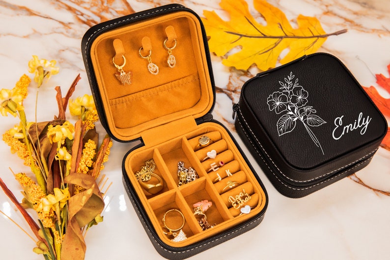 Puede incluir: Caja de joyer&iacute;a de cuero negro con un dise&ntilde;o floral y el nombre "Emily" grabado en la tapa. La caja est&aacute; abierta, revelando un interior de terciopelo amarillo con compartimentos para anillos, aretes y otras joyas.