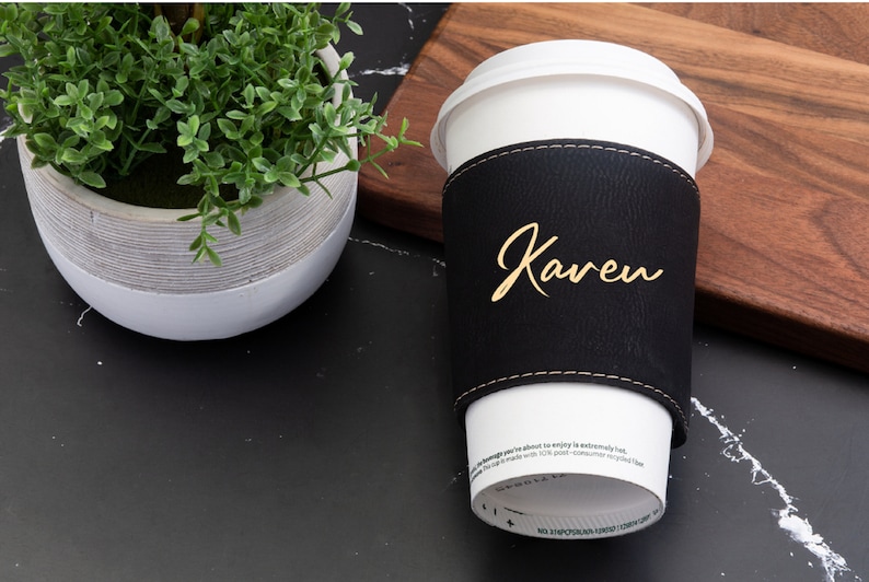 Puede incluir: Una funda de taza de caf&eacute; de cuero negro con el nombre "Karen" escrito en letras doradas. La funda est&aacute; en una taza de caf&eacute; blanca.