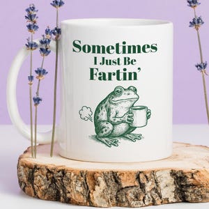 May include: White ceramic mug with the text "Sometimes I Just Be Fartin'" in green. A green frog illustration holding a mug is also on the mug. The mug is on a wooden surface with lavender sprigs.