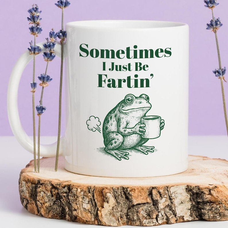 May include: White ceramic mug with the text "Sometimes I Just Be Fartin'" in green. A green frog illustration holding a mug is also on the mug. The mug is on a wooden surface with lavender sprigs.