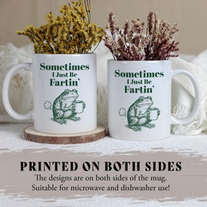 May include: Two white ceramic mugs with a humorous frog design. Each mug features the text "Sometimes I Just Be Fartin'" and a green illustration of a frog holding a cup. The mugs are filled with dried flowers and sit on wooden coasters.