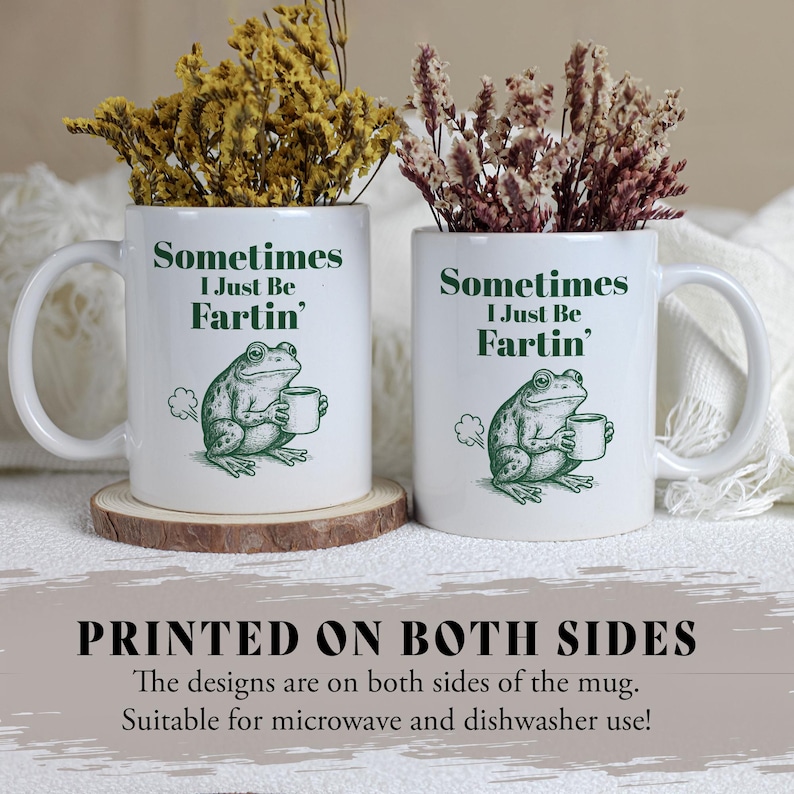 May include: Two white ceramic mugs with a humorous frog design. Each mug features the text "Sometimes I Just Be Fartin'" and a green illustration of a frog holding a cup. The mugs are filled with dried flowers and sit on wooden coasters.