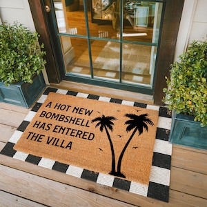 May include: A rectangular tan coir doormat featuring the black text A HOT NEW BOMBSHELL HAS ENTERED THE VILLA, accompanied by black silhouettes of two palm trees and several small birds, placed on a black and white striped rug.