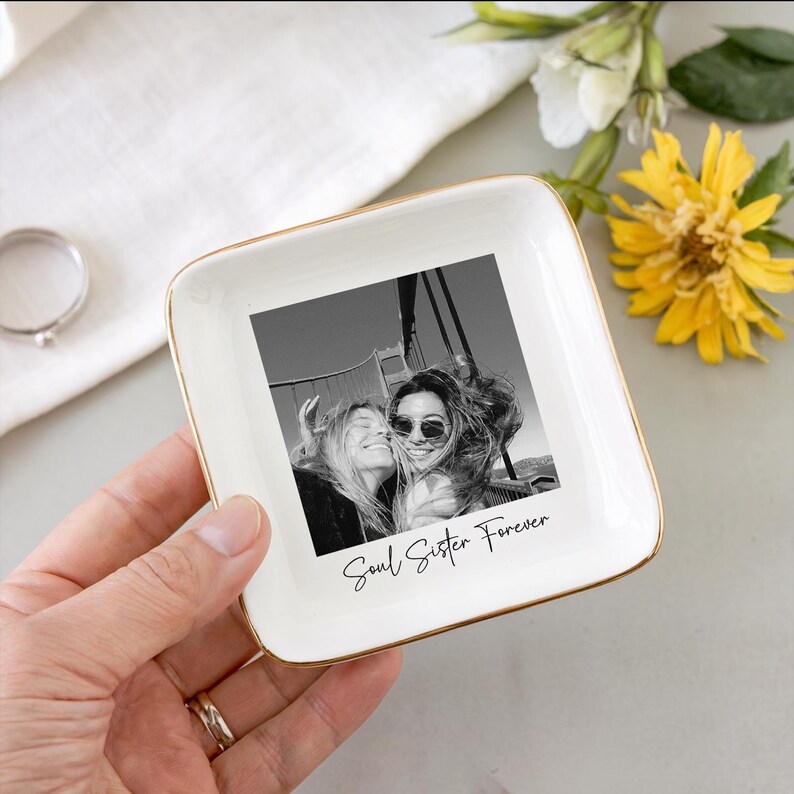 May include: A white ceramic trinket dish with a gold rim, featuring a black and white photo of two people. The words "Soul Sister Forever" are written below the photo. The dish is held by a hand.