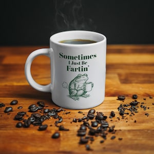 May include: White ceramic coffee mug with the text "Sometimes I Just Be Fartin'" and a green illustration of a frog holding a coffee cup. The mug is filled with dark coffee, and coffee beans are scattered around it.