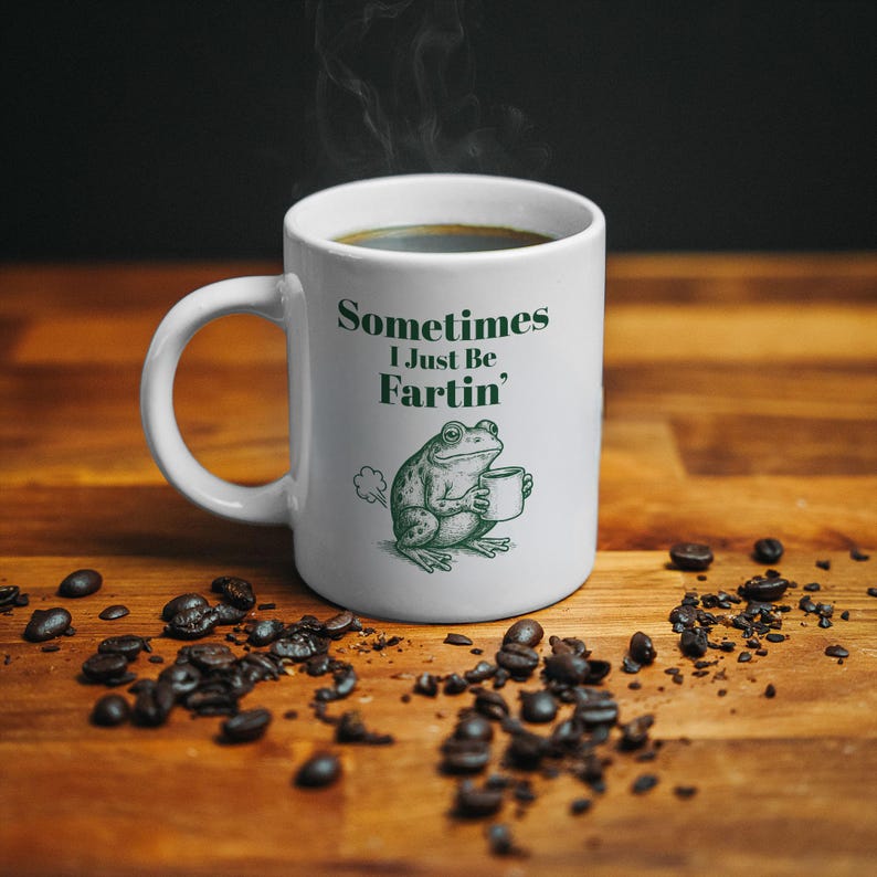 May include: White ceramic coffee mug with the text "Sometimes I Just Be Fartin'" and a green illustration of a frog holding a coffee cup. The mug is filled with dark coffee, and coffee beans are scattered around it.