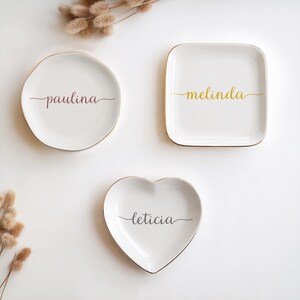 May include: Three white ceramic trinket dishes with gold trim. The dishes are shaped like a circle, a square, and a heart. The names "Paulina", "Melinda", and "Leticia" are written in cursive on each dish.