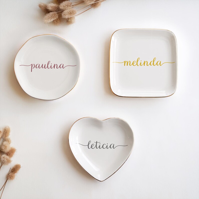 May include: Three white ceramic trinket dishes with gold trim. The dishes are shaped like a circle, a square, and a heart. The names "Paulina", "Melinda", and "Leticia" are written in cursive on each dish.