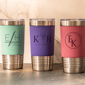 May include: Three stainless steel tumblers with personalized leather wraps. The tumblers are in the colors teal, purple, and pink. The teal tumbler has the initials "E/H" and the name "EMIL HUTCHINSON". The purple tumbler has the initials "KVR" and the name "KATE ROBERTS". The pink tumbler has the initials "EK" and the name "ELIZA KATHMAN".